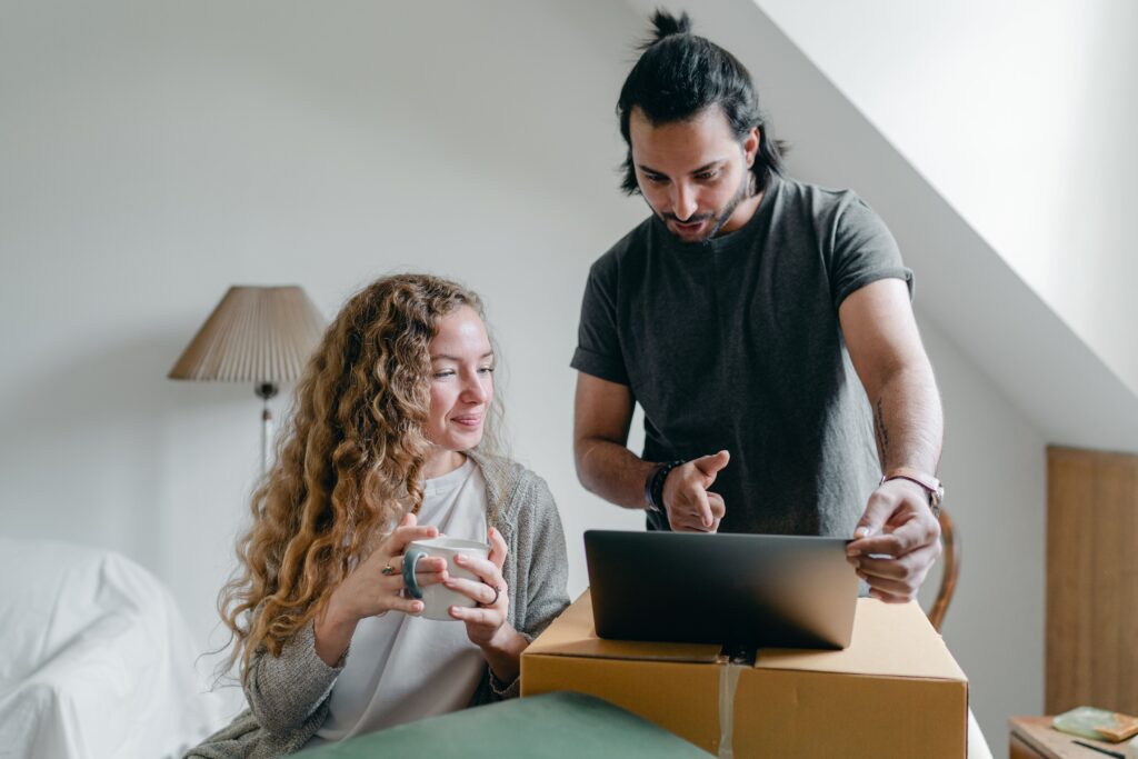 Positive happy young couple in casual clothes packing belongings into carton box and using laptop in living room near window